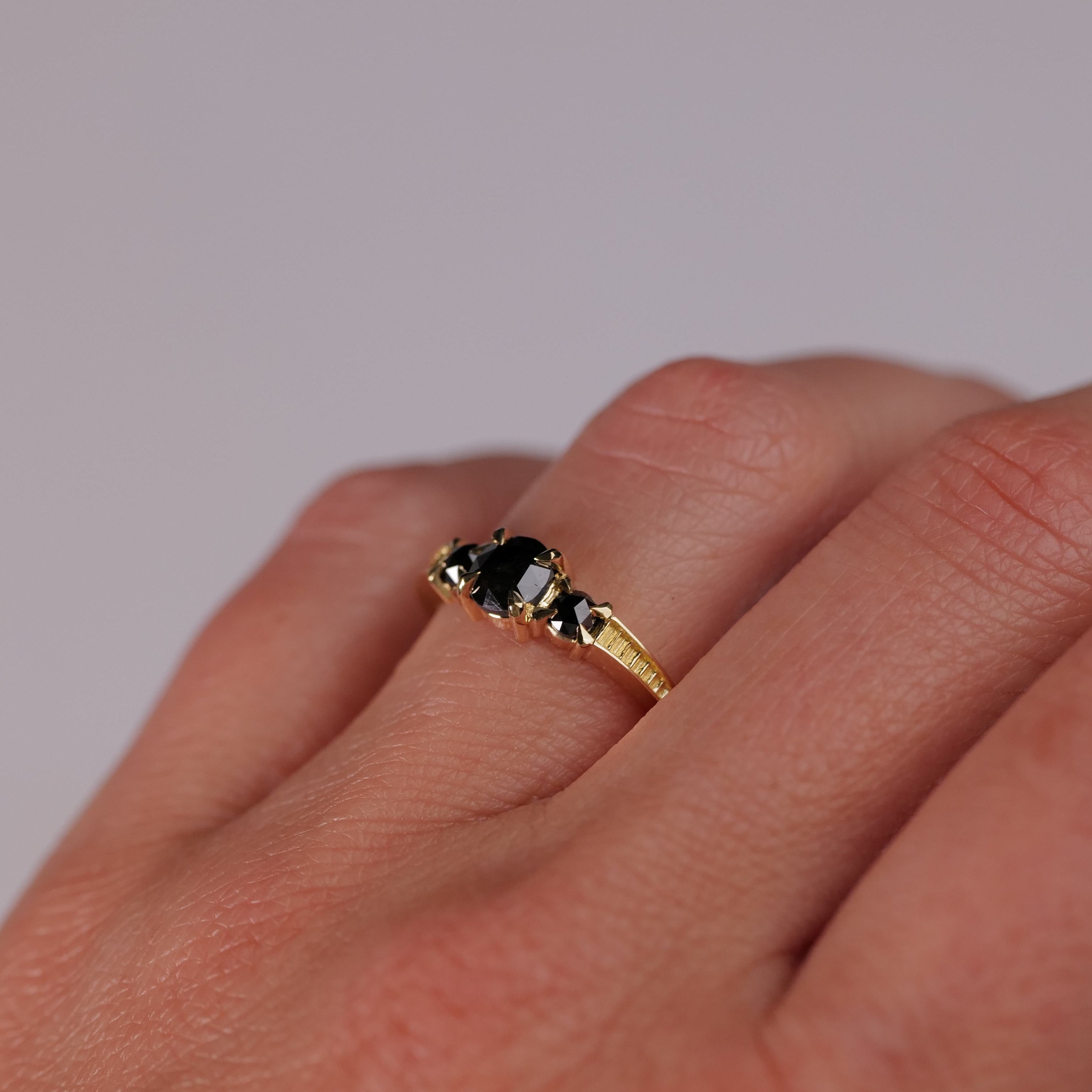 A Fiat Lux Classic 3 Stone Temple Ring - Black Diamond shines on a hand with a soft, neutral background.