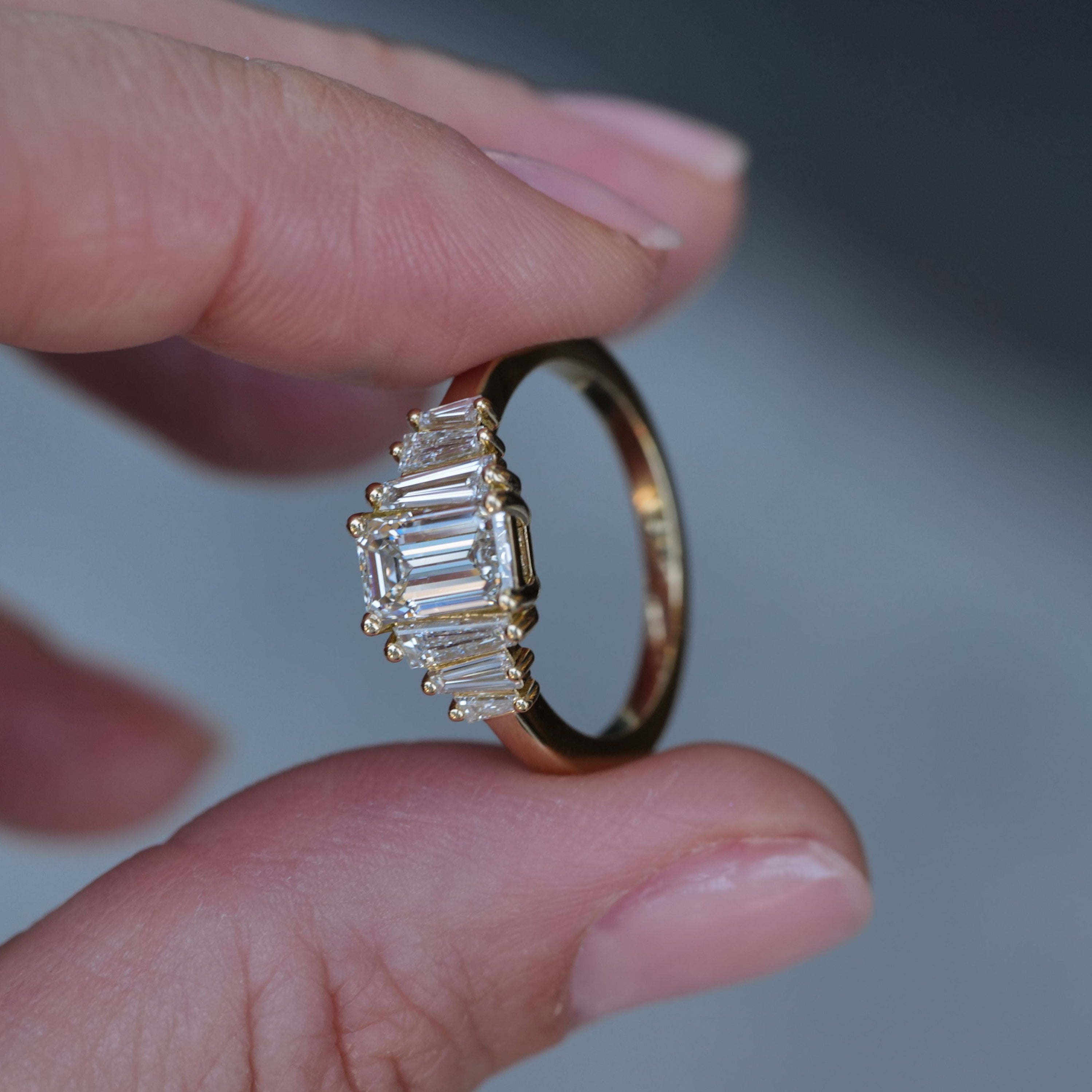A hand holds Fiat Luxs Classic Calypso ring with a 1.02ct emerald cut diamond and side stones.