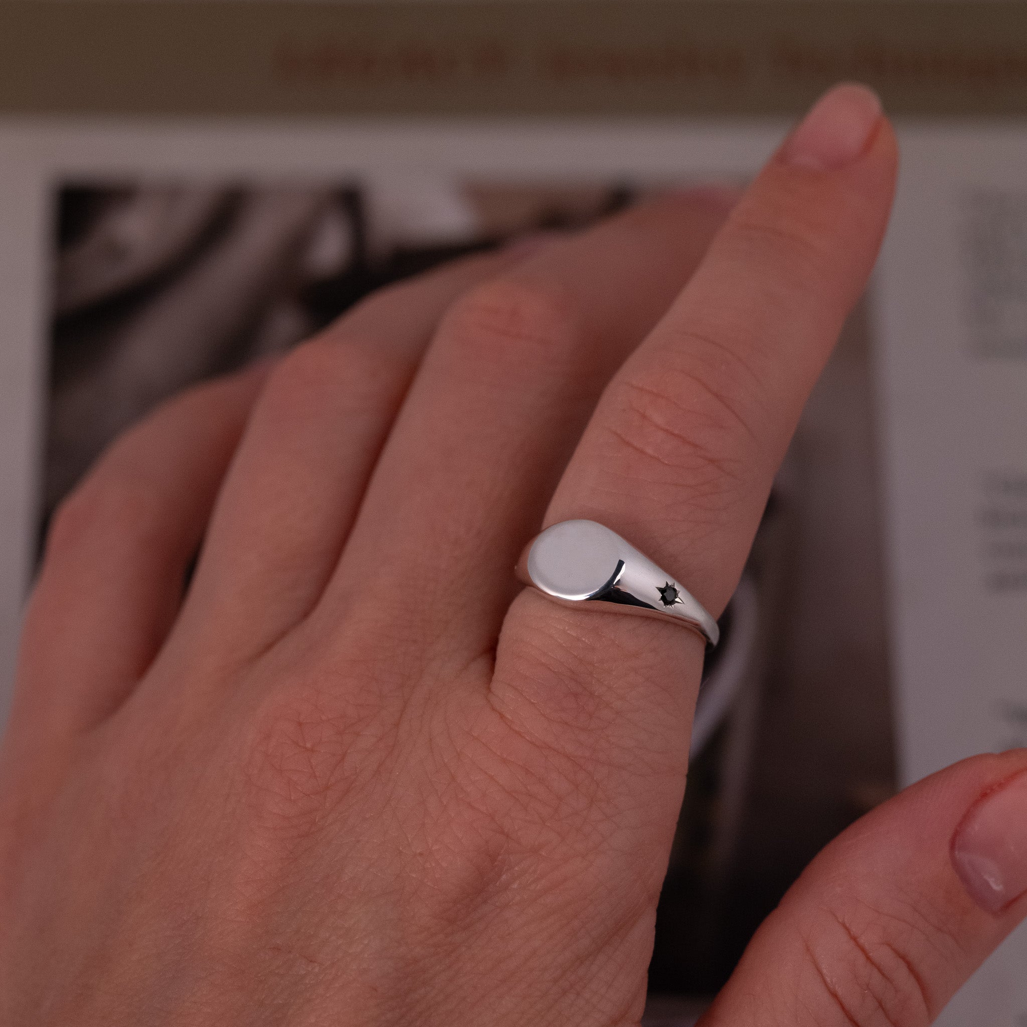A hand wears the Fiat Lux Classic Horizontal Oval Signet Ring - Star Set (S) on the pinky, book blurred in background.