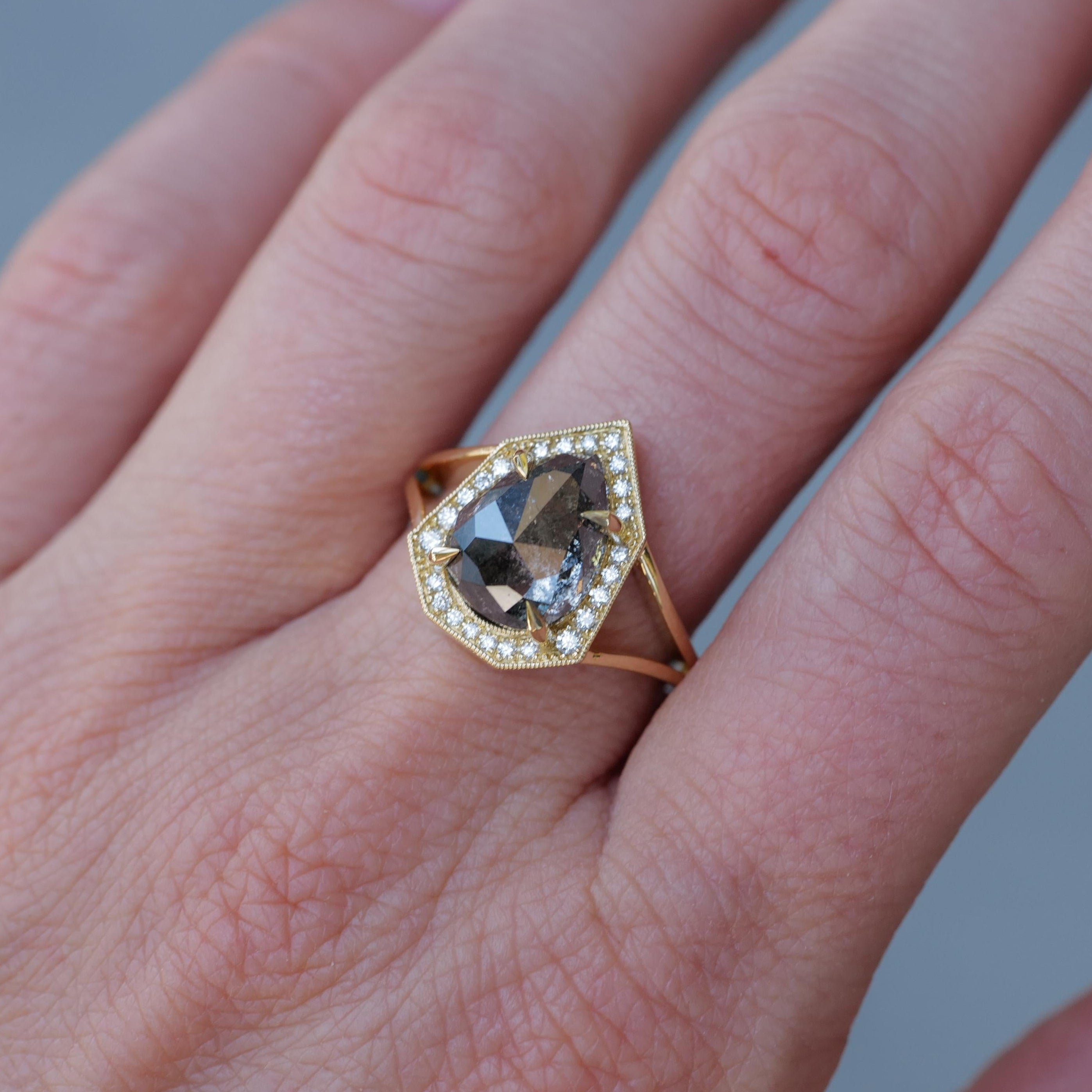 A hand wears Fiat Lux’s Grande Phoebe ring: a 4.88ct salt & pepper diamond framed by small white diamonds in 18K gold.