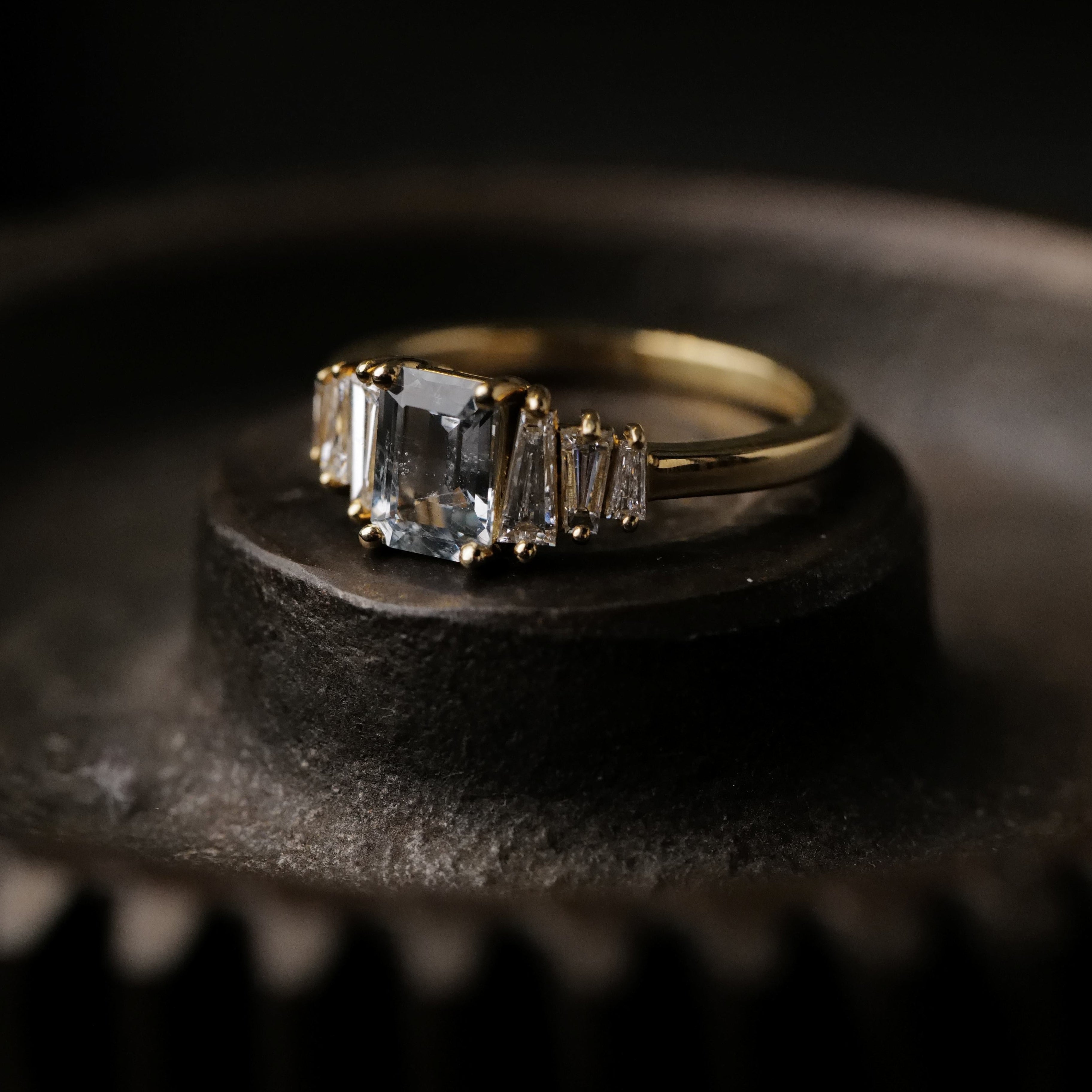 The Fiat Lux Petite Calypso ring with a 1.16ct ice blue sapphire rests on a dark, ridged surface.
