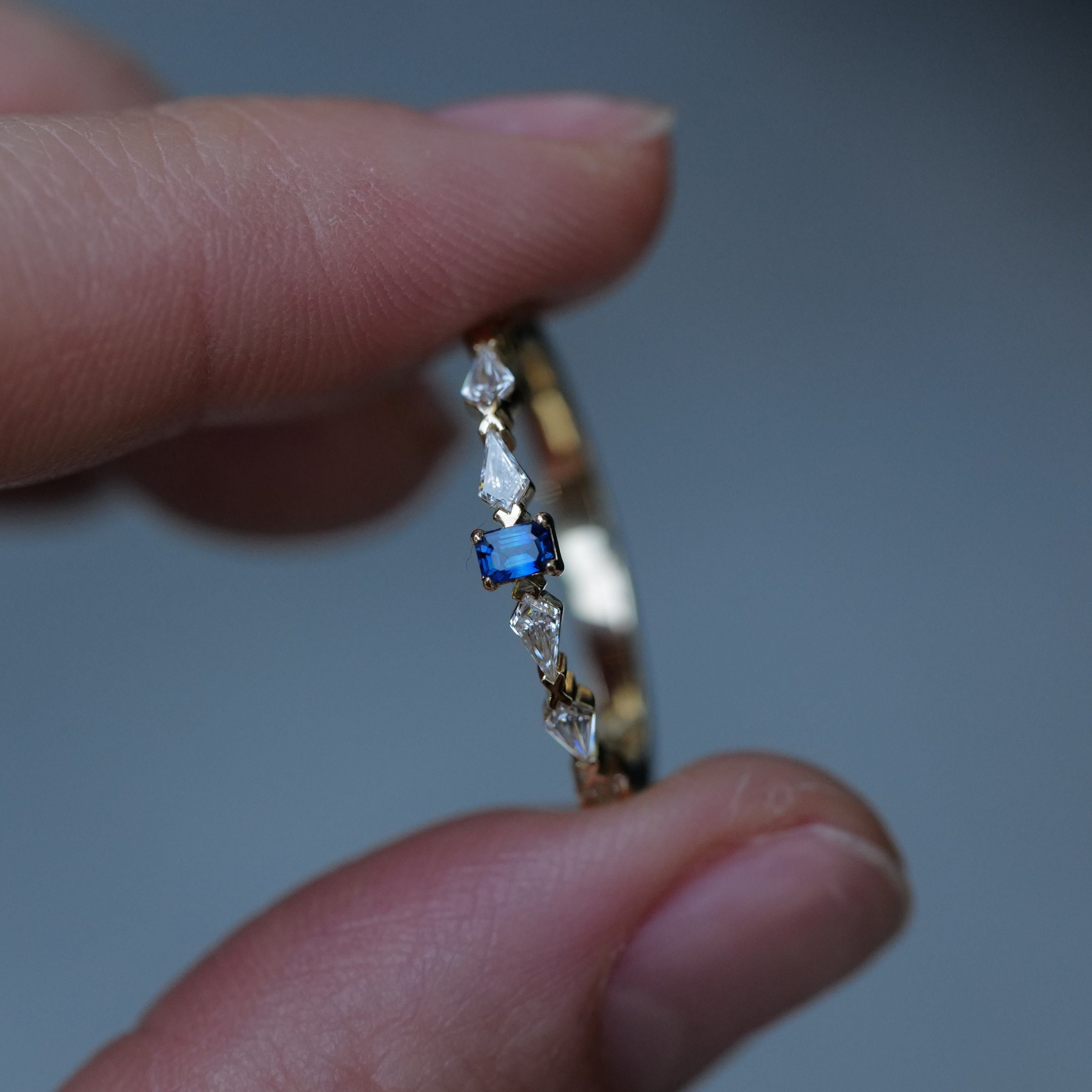 A hand holds the Fiat Lux Petite Astra Ring in 14K gold with a blue rectangular gem and kite-shaped diamonds.