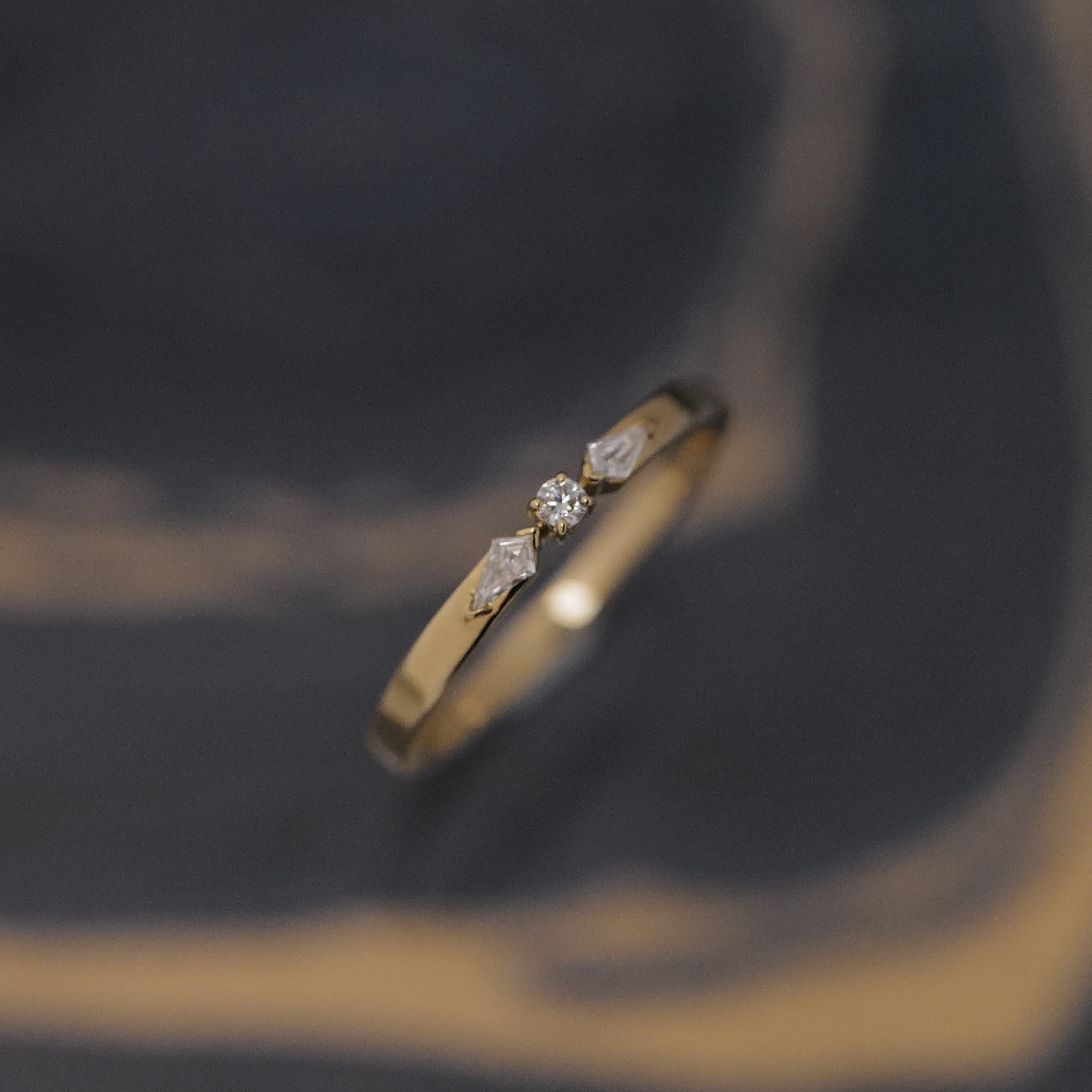 The Fiat Lux Petite Nova Ring features a small central diamond with geometric accents on a blurred dark background.