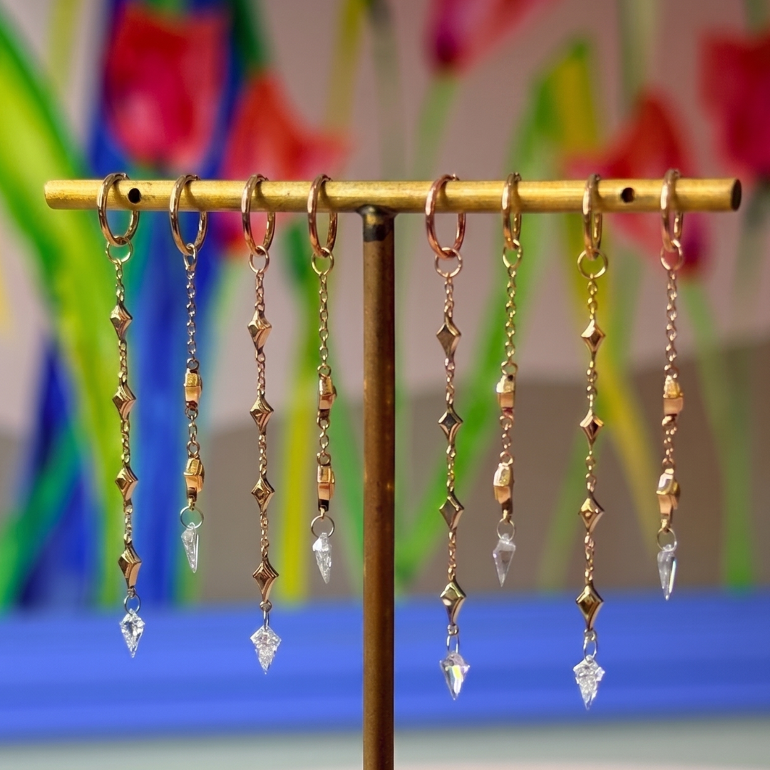 Fiat Lux Constellation Kite Dangle: 14K gold earrings with beads & crystals, shown on brass stand with floral backdrop.