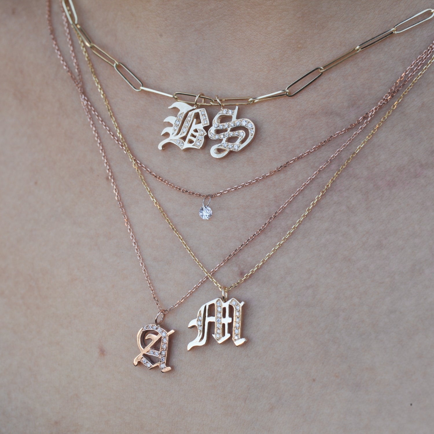 Four layered necklaces: paperclip chain, Georgie Gold Ideogram Charm, solitaire gem, and A & M pendants on a neck.