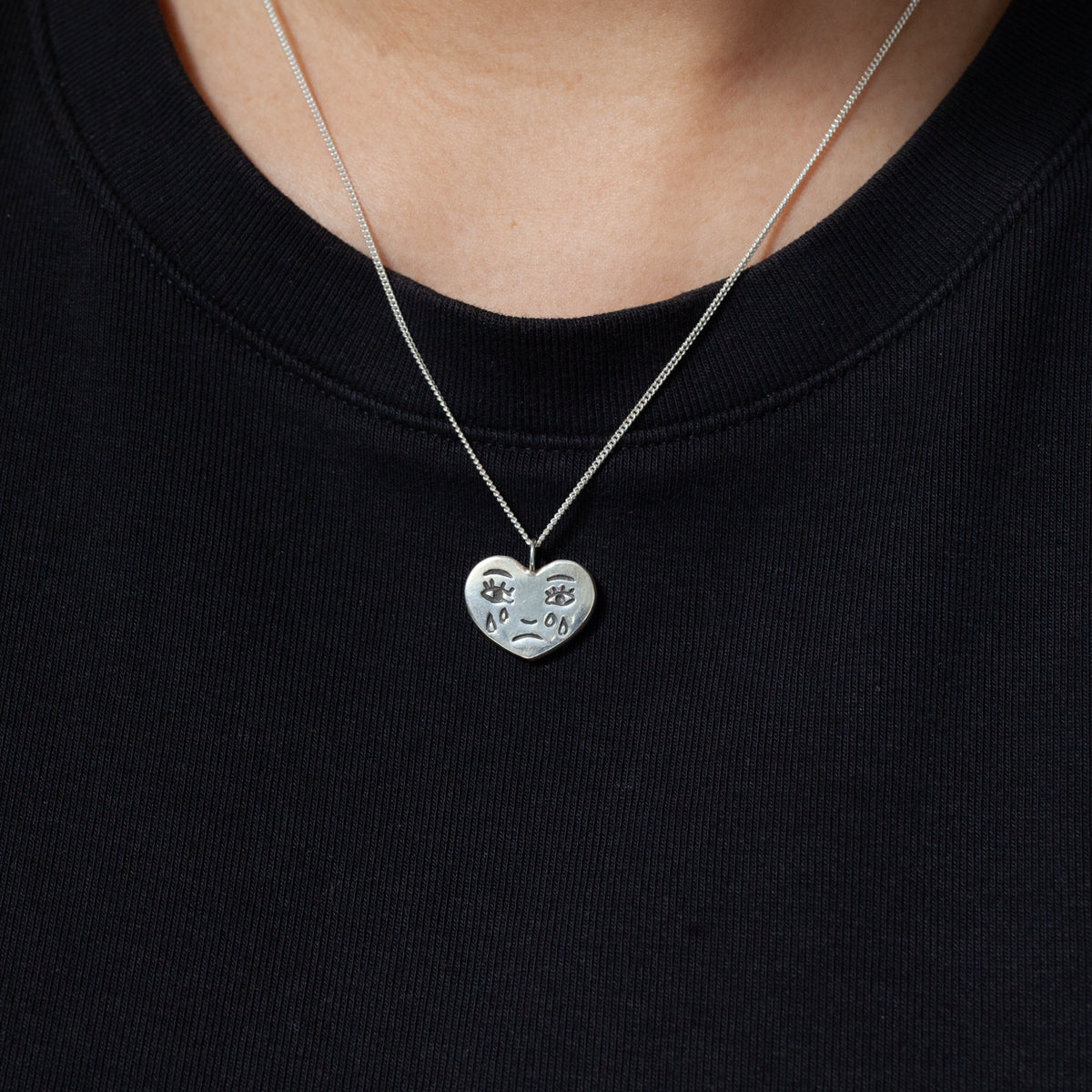 The Ali Munn Grimm Necklace features a sad face pendant on silver chain, styled over a black crew-neck shirt.