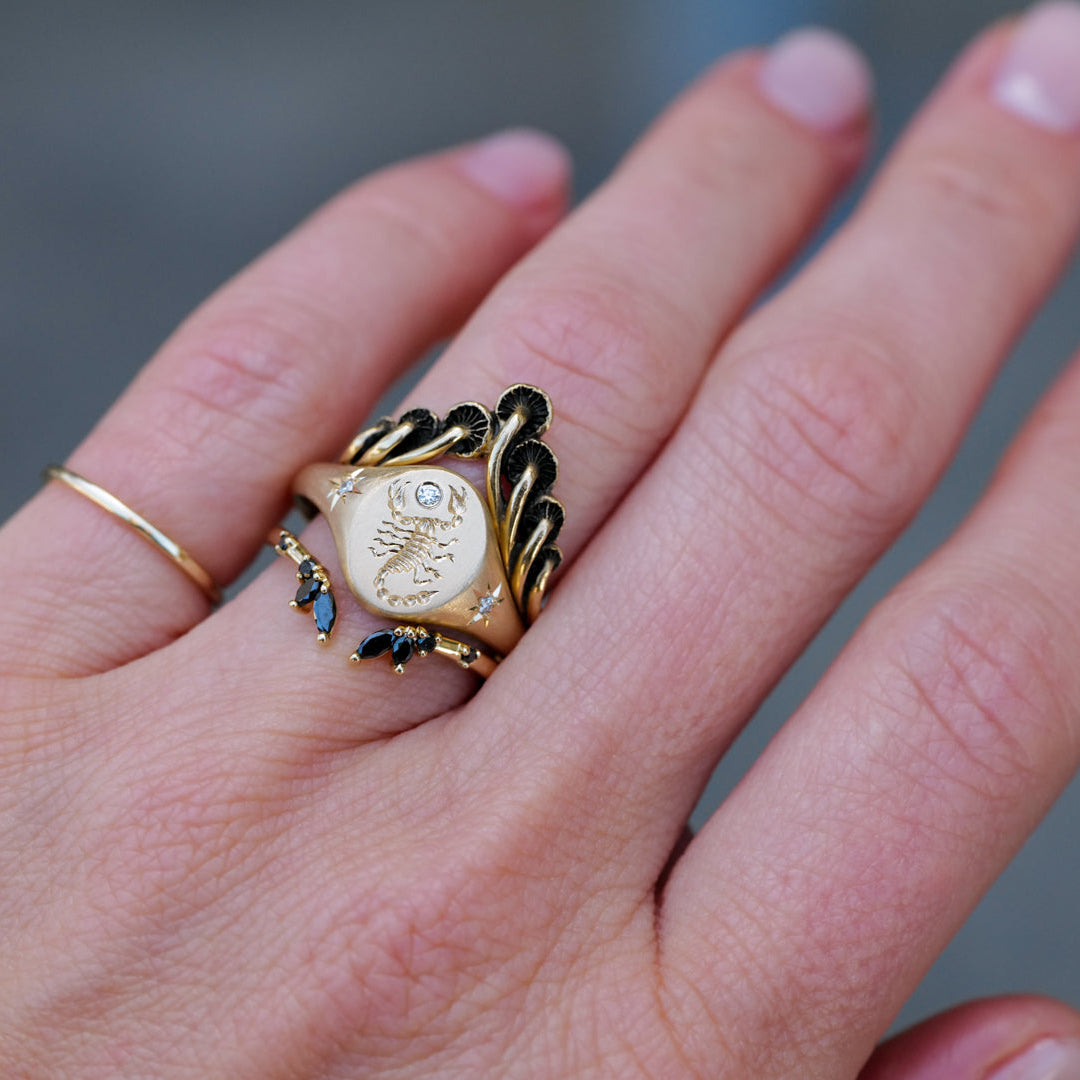 A hand wears three 14k gold rings, including Blue Blue Driver’s Scorpion Signet Ring - Diamond and a sun-engraved signet.