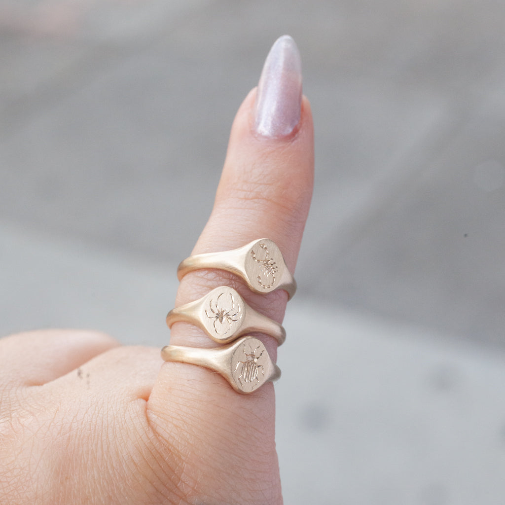A hand with metallic nails wears three 14K gold signet rings, one is Blue Blue Drivers Tiniest Scarab Signet Ring on the thumb.
