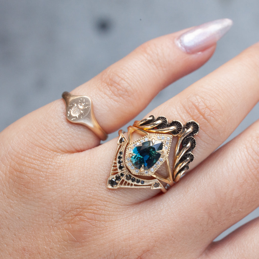 A hand with a 14K gold signet ring and Blue Blue Drivers Tiniest Scarab Signet Ring in gold with teal and black details.