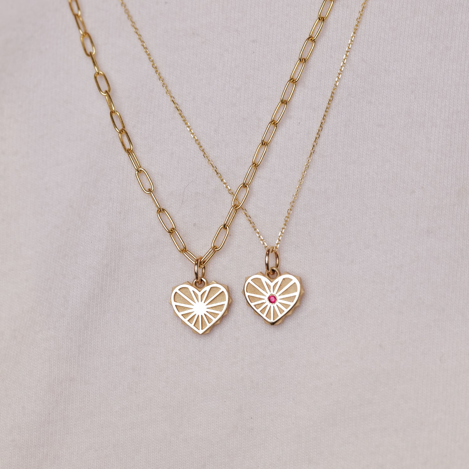 Two EMBLM Twin Flame Heart Charm gold necklaces, one with a pink stone, displayed on light fabric.
