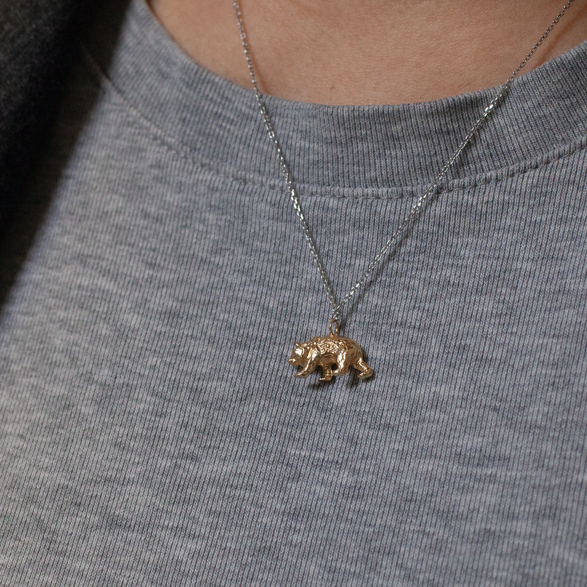 A small 10k gold Bear Charm by Fiat Lux on a delicate silver chain worn around a person’s neck.