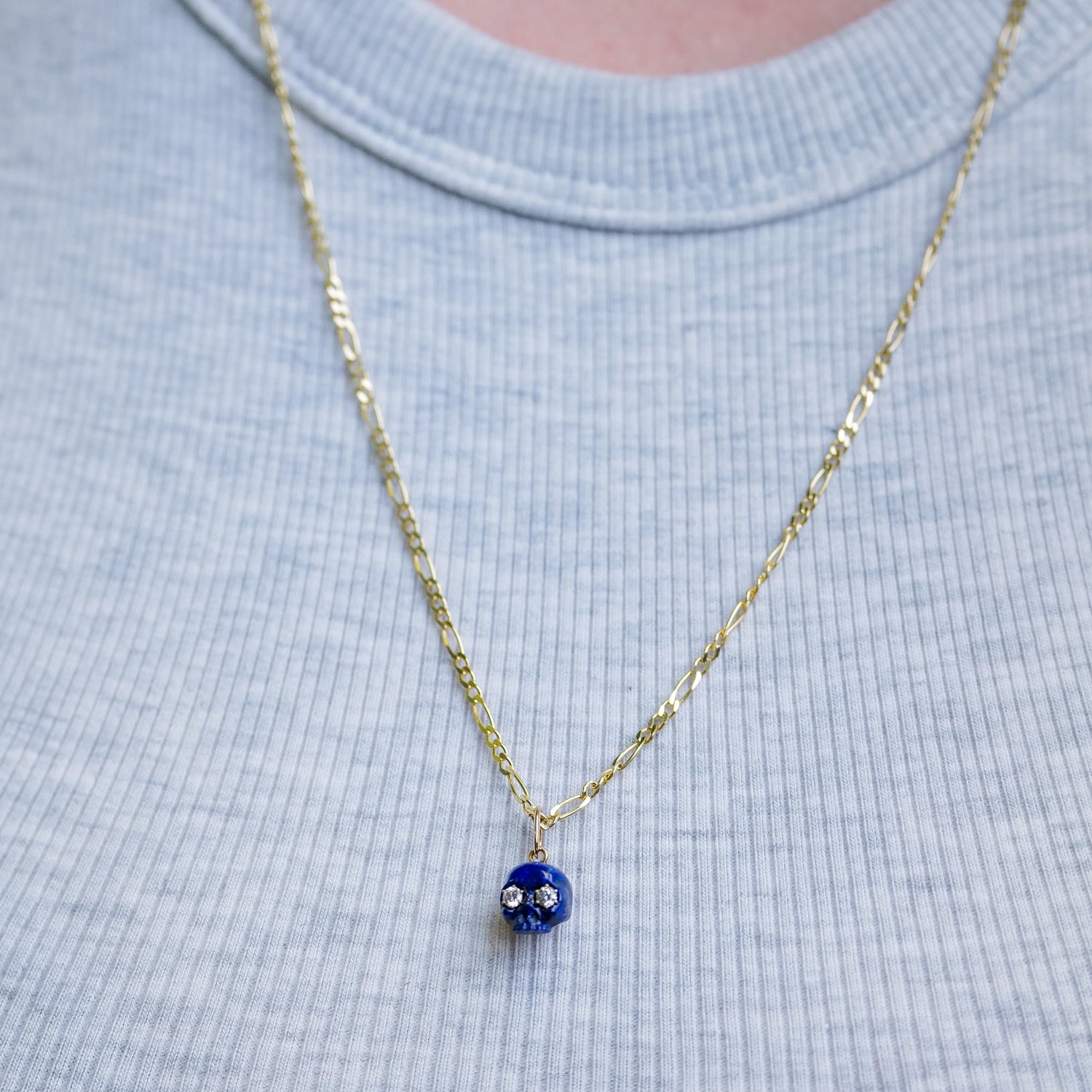 Fiat Luxs The Skull of Lapis: a 14K gold chain necklace with blue bead pendant, white flowers, on light blue shirt.