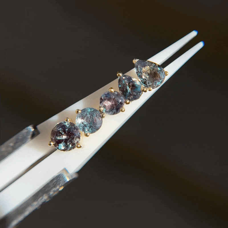 Five pear-cut flatback Alexandrite 3 Prong Pear Grande gemstones in a gold setting, held by white tweezers.