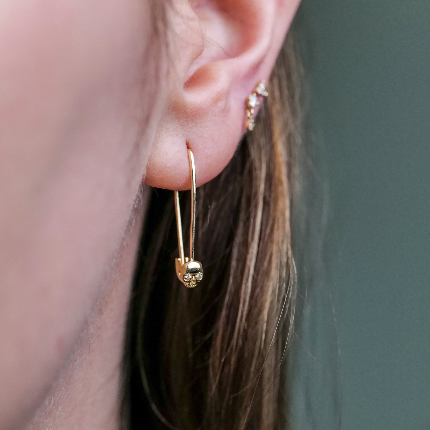 Close-up of an ear wearing Fiat Luxs Skull Safety Pin 14k gold earring and a small stud in the upper ear.