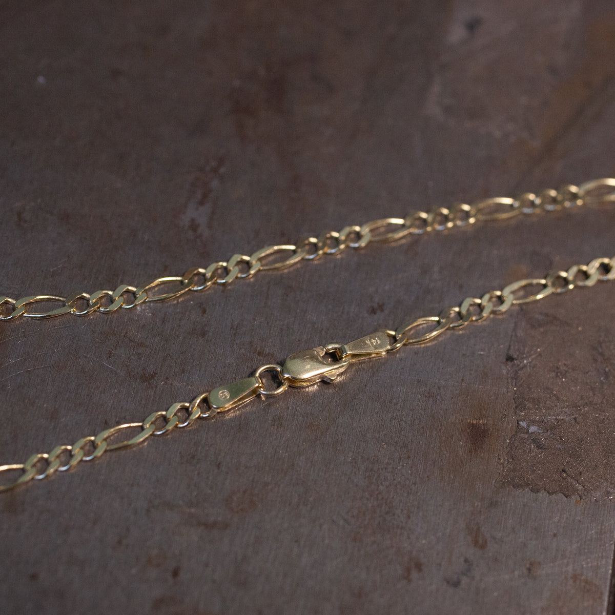 Two Fiat Lux Classic Figaro Chains in 14K gold with lobster clasps rest on a textured brown surface.