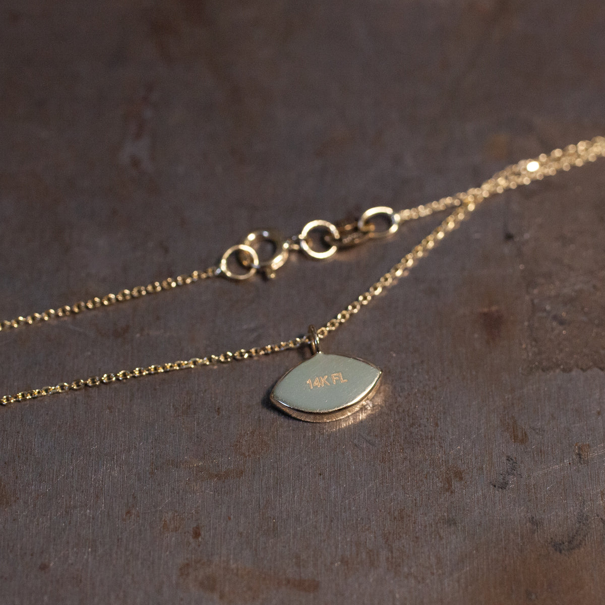 Fiat Lux Mini Mati Necklace (G): a delicate gold necklace with a small 14K FL pendant on a brown surface.