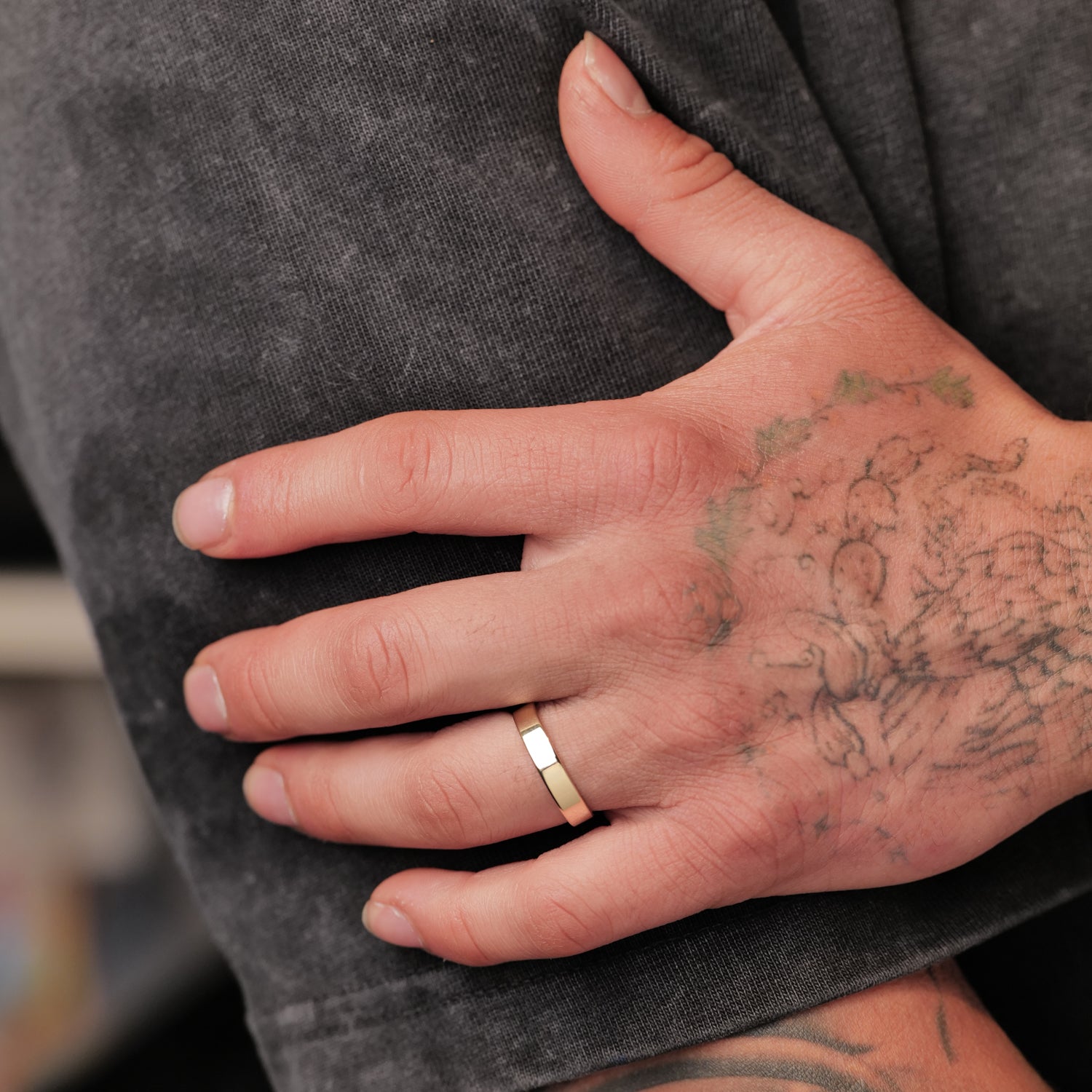 A tattooed hand with a Fiat Lux Classic Flat Band (4mm) rests on a person’s leg in gray pants.