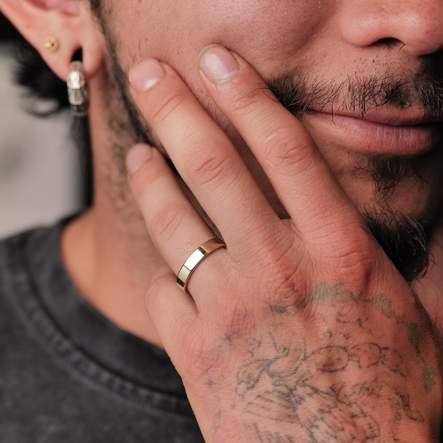 A mustachioed person with hand tattoos touches their face, wearing Fiat Lux’s 4mm Classic Flat Band and gold earrings.