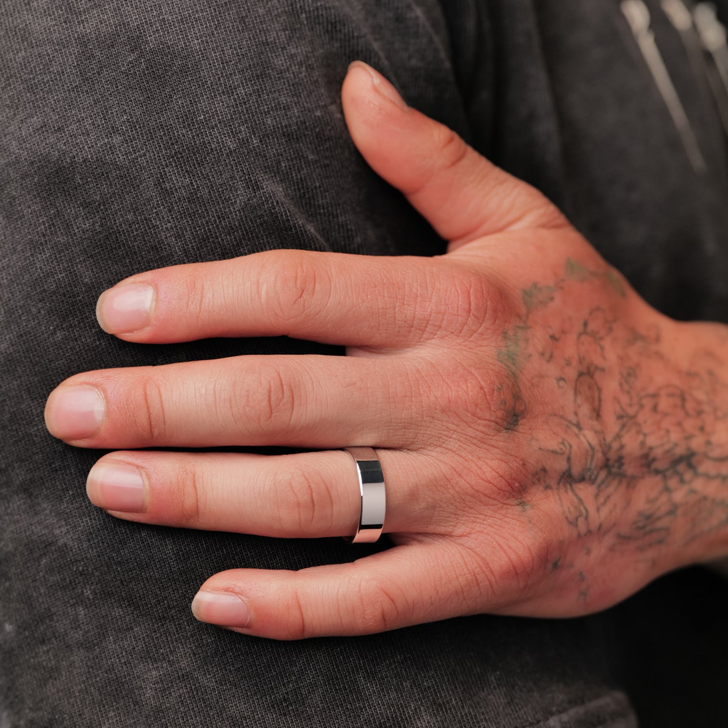 A tattooed hand rests on a shoulder, wearing the Fiat Lux Classic Flat Band (6mm) on the ring finger.
