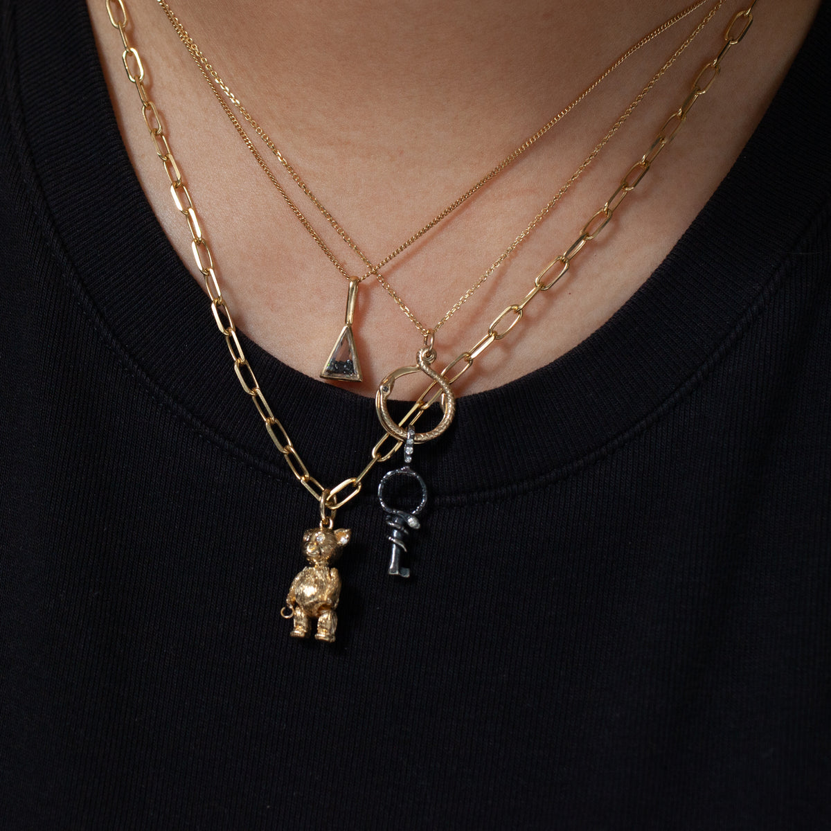 Moritz Glik Triangle Shaker Charm: 18K gold, worn layered with hoop and bear-key necklaces over a black shirt.