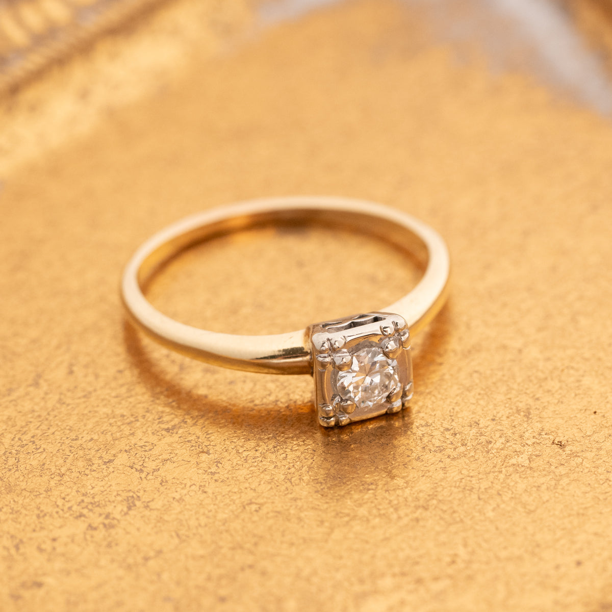 A vintage gold solitaire engagement ring with a square-cut clear gemstone in a four-prong setting, resting on a textured gold surface.