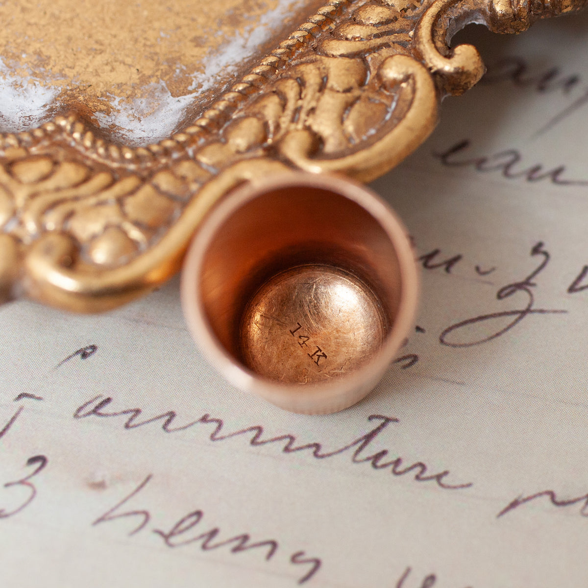 A Vintage Engraved Thimble in 14k gold with a plant motif, resting on a handwritten document next to an ornate tray.