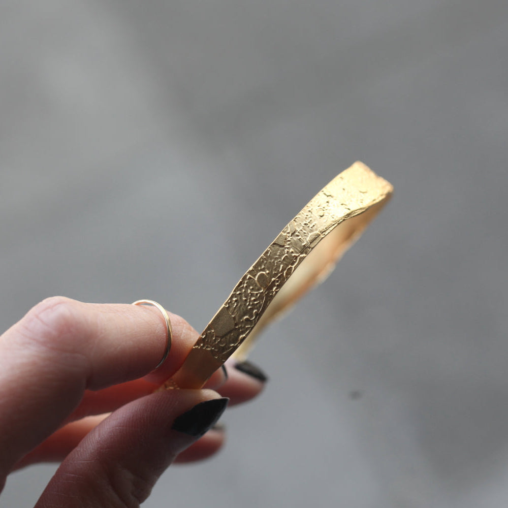 A hand holds Lingua Nigras Acid-Etched Bangle, a unique 22k gold bracelet, against a blurred gray background.