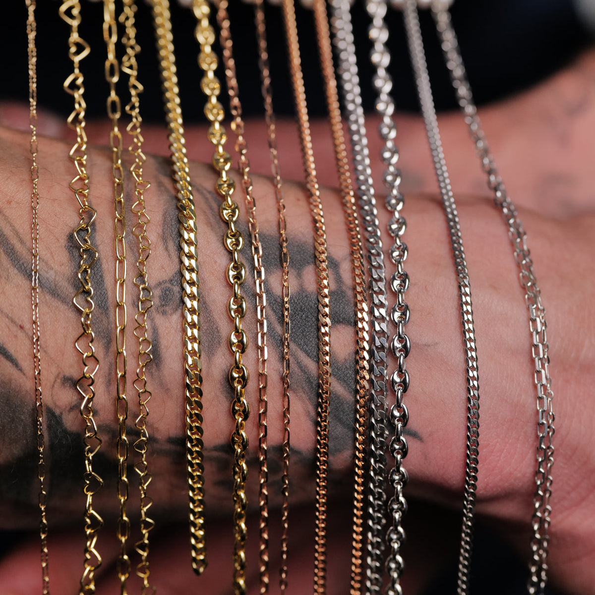 A variety of gold, rose gold, and silver metal chains draped over a tattooed forearm.