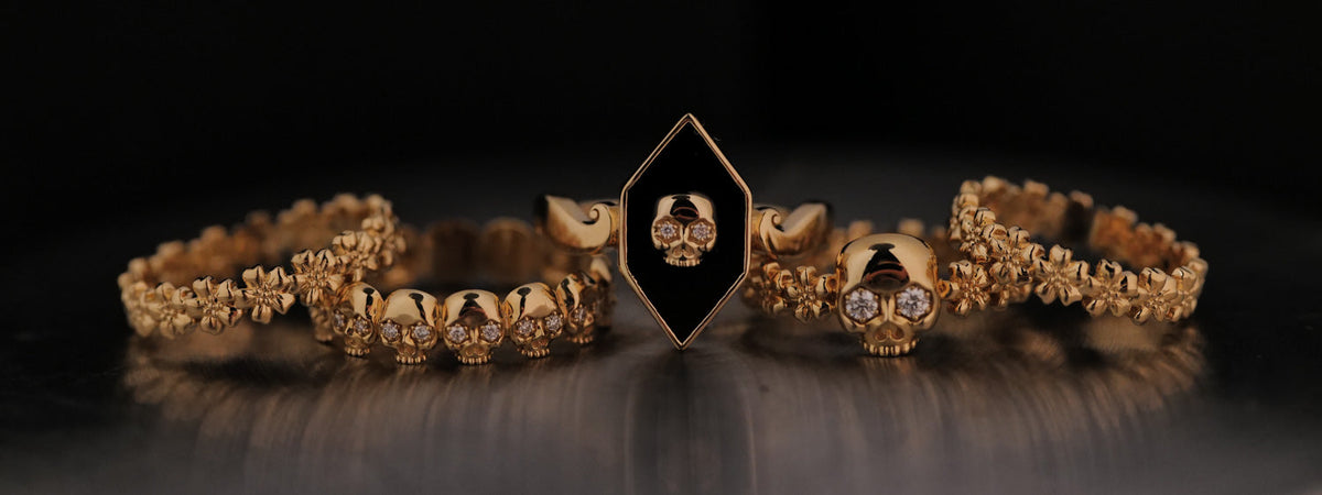 Three gold rings: one with a diamond-shaped black inlay featuring a skull, one with a row of small skulls, and one featuring a single skull flanked by floral bands.