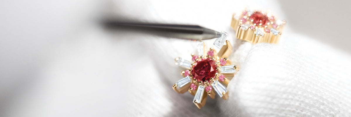 Gold stud earrings featuring a central red gemstone surrounded by pink stones and clear baguette-cut accents, held by a pair of tweezers.