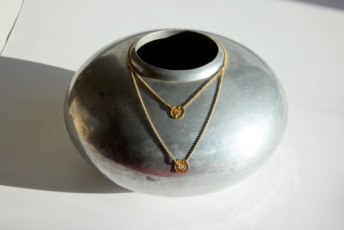 A gold layered necklace with two circular pendants draped over a smooth, round metallic vessel.