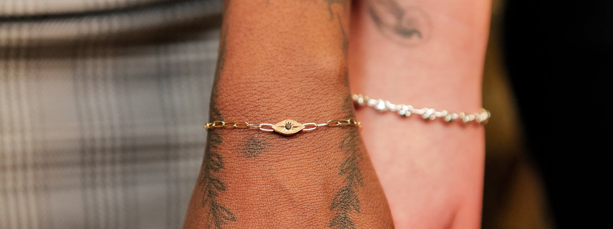 Gold chain bracelet with an evil eye charm featuring a small black stone at the center, worn on a tattooed wrist.