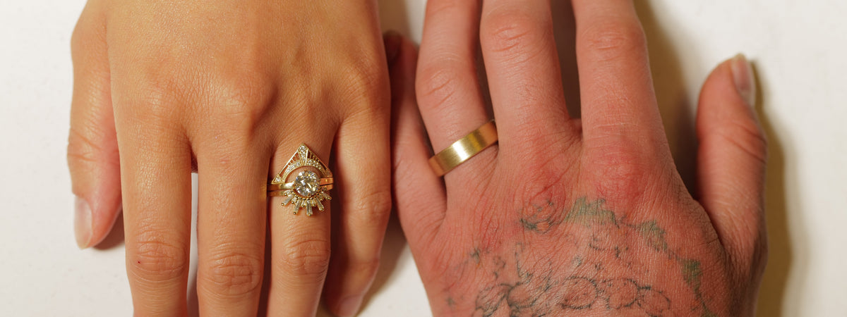 Two hands side by side, one wearing a decorative gold diamond ring set and the other wearing a plain gold band.