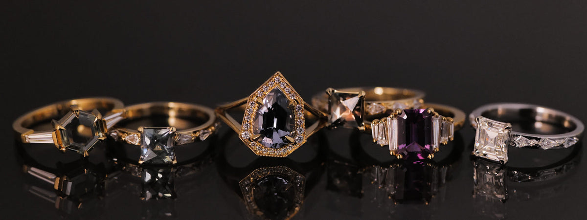 A row of six assorted gemstone rings, including diamond, pear-shaped, and rectangular-cut stones, set in gold and silver bands on a reflective black surface.