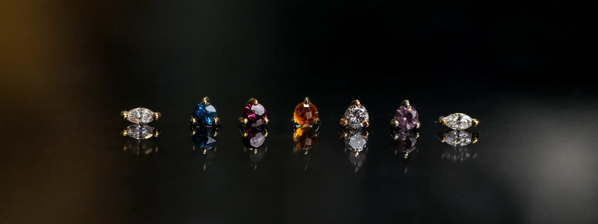 Seven individual gemstone stud earrings in various colors and shapes, arranged in a row against a black reflective background.
