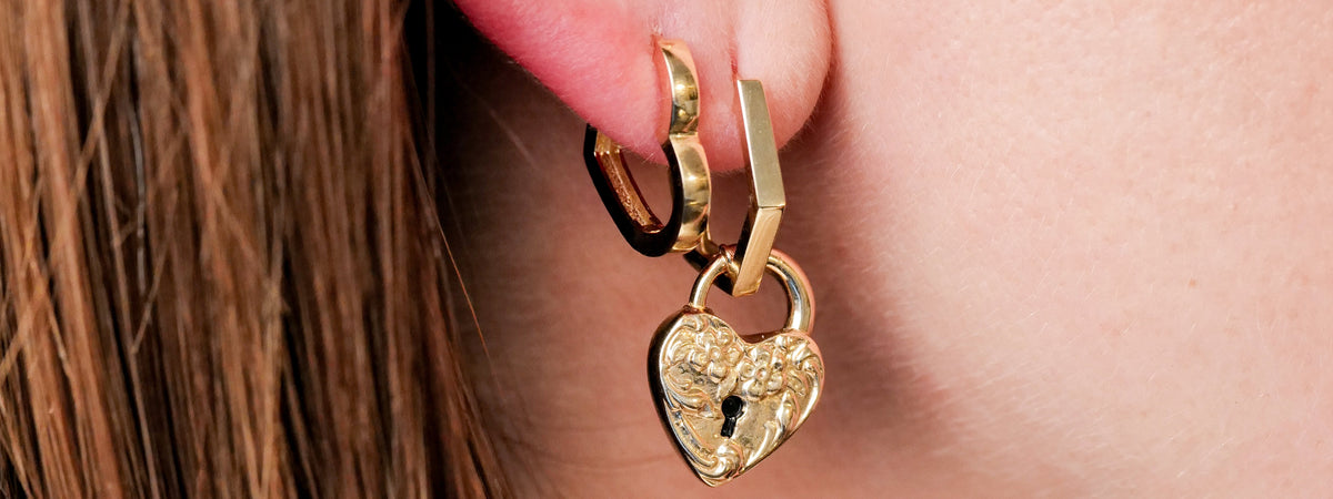 Gold hoop earring with a heart-shaped padlock charm hanging from the earlobe.