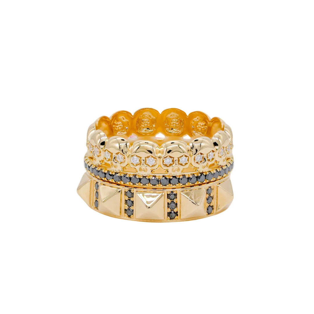 Three stacked gold rings, including Fiat Lux’s Eternity Skull Ring in 14k gold with black stones, on white background.
