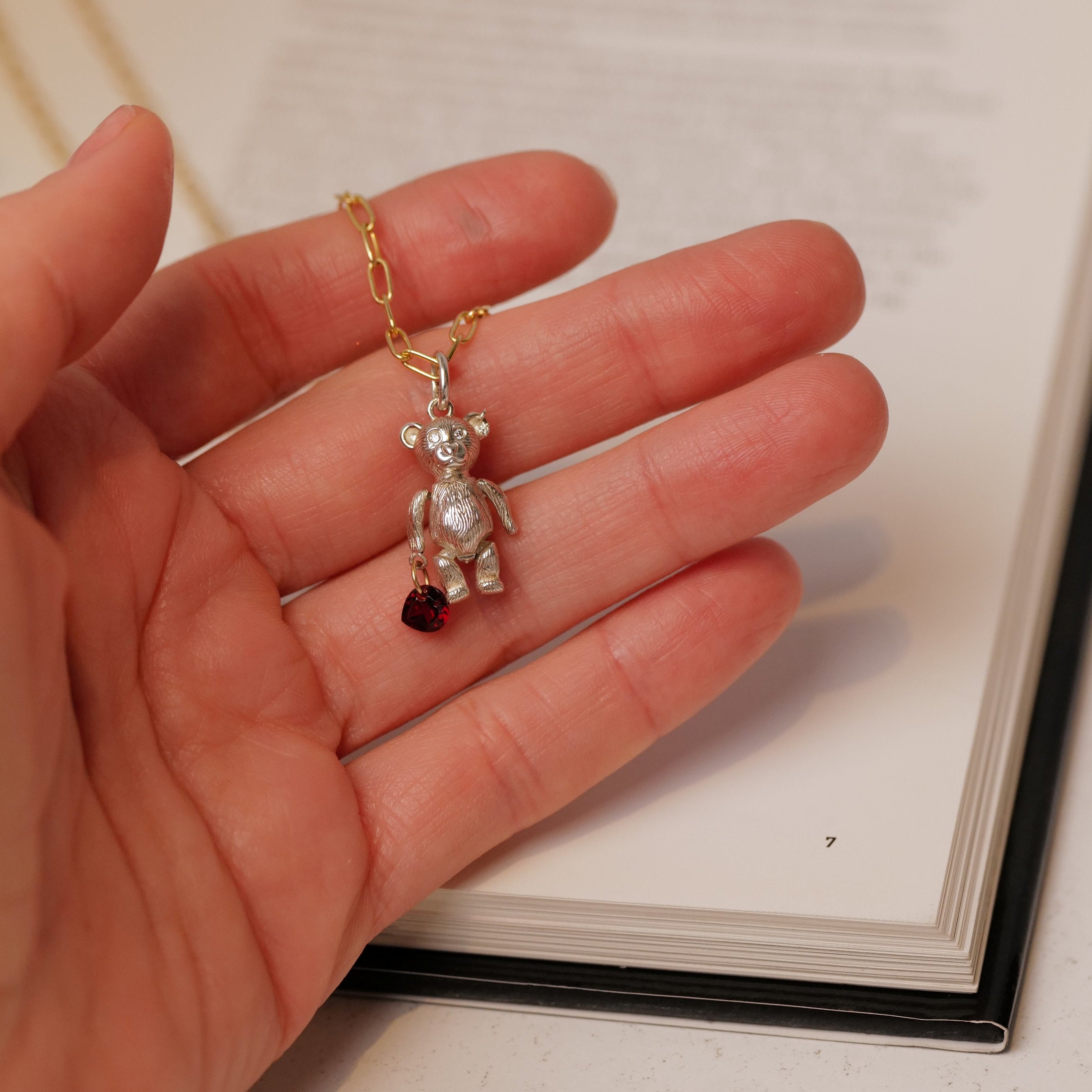 A hand holds the Fiat Lux Mr Teddy Charm (S) necklace with a small red heart, above an open book.