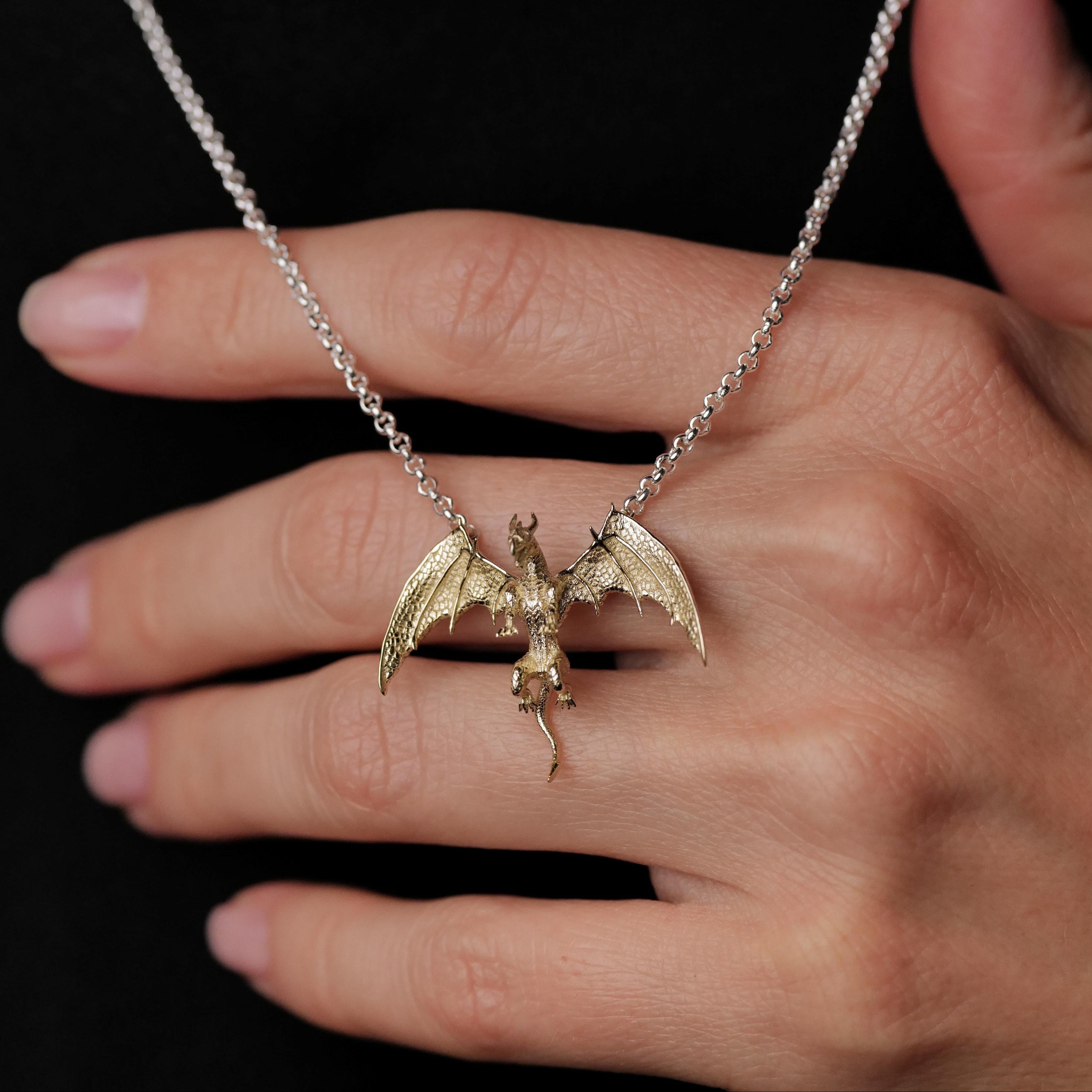 A hand holds the Fiat Lux Medieval Dragon Necklace (G), featuring a 10K gold dragon charm with outstretched wings.