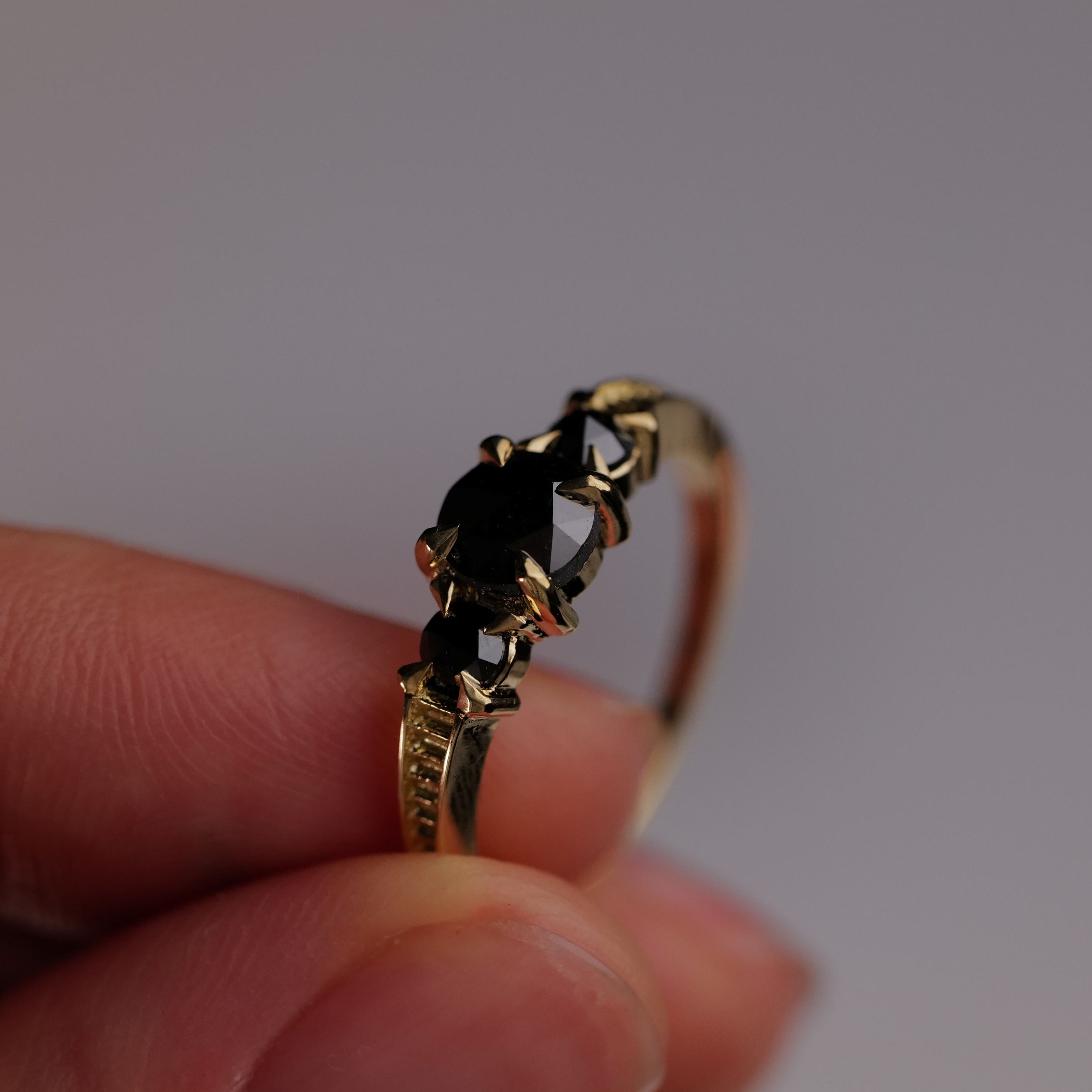 A hand holds Fiat Luxs Classic 3 Stone Temple Ring - Black Diamond against a plain, light background.