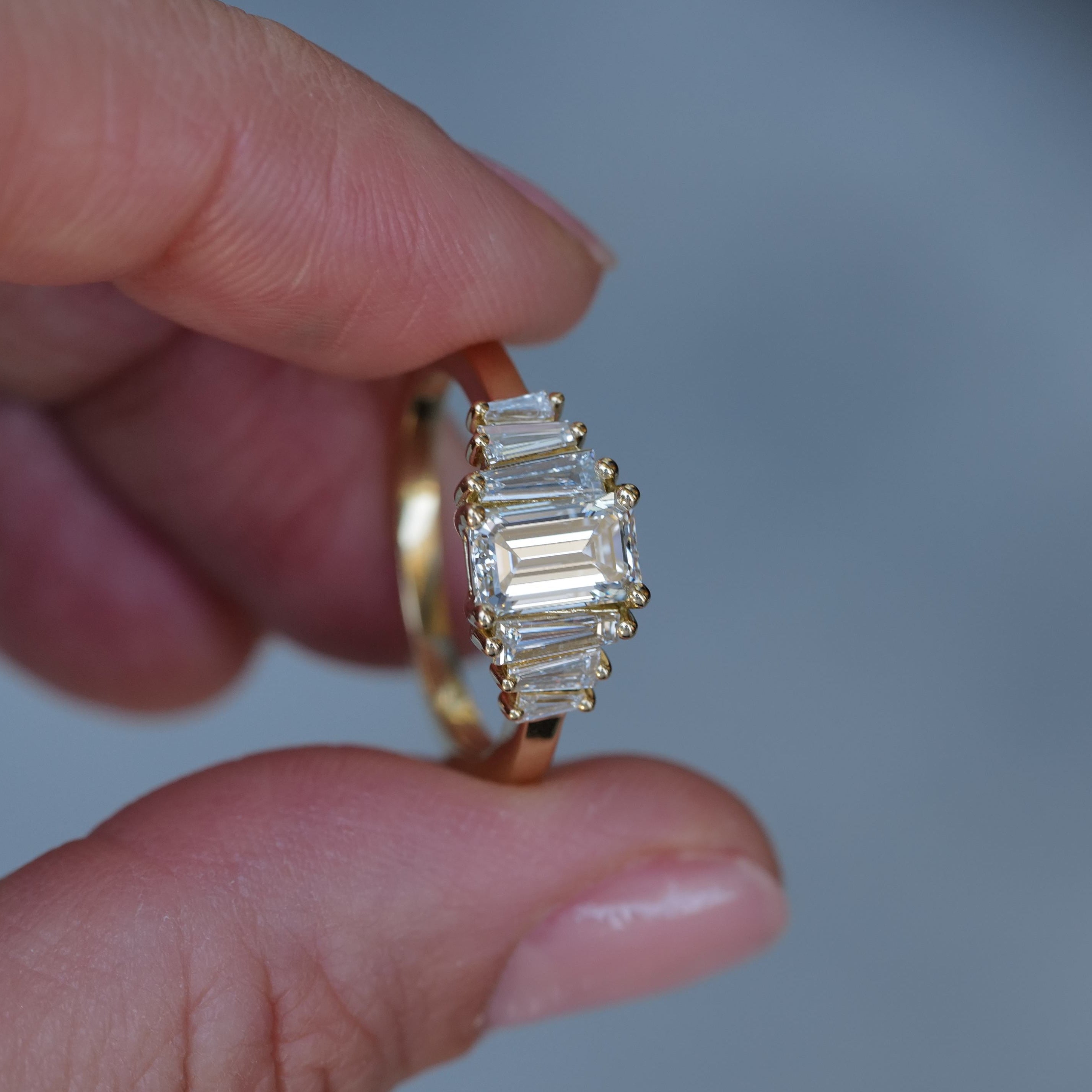 A hand holds the Fiat Lux Classic Calypso ring, featuring 1.02ct emerald cut diamonds in a sleek gold band.