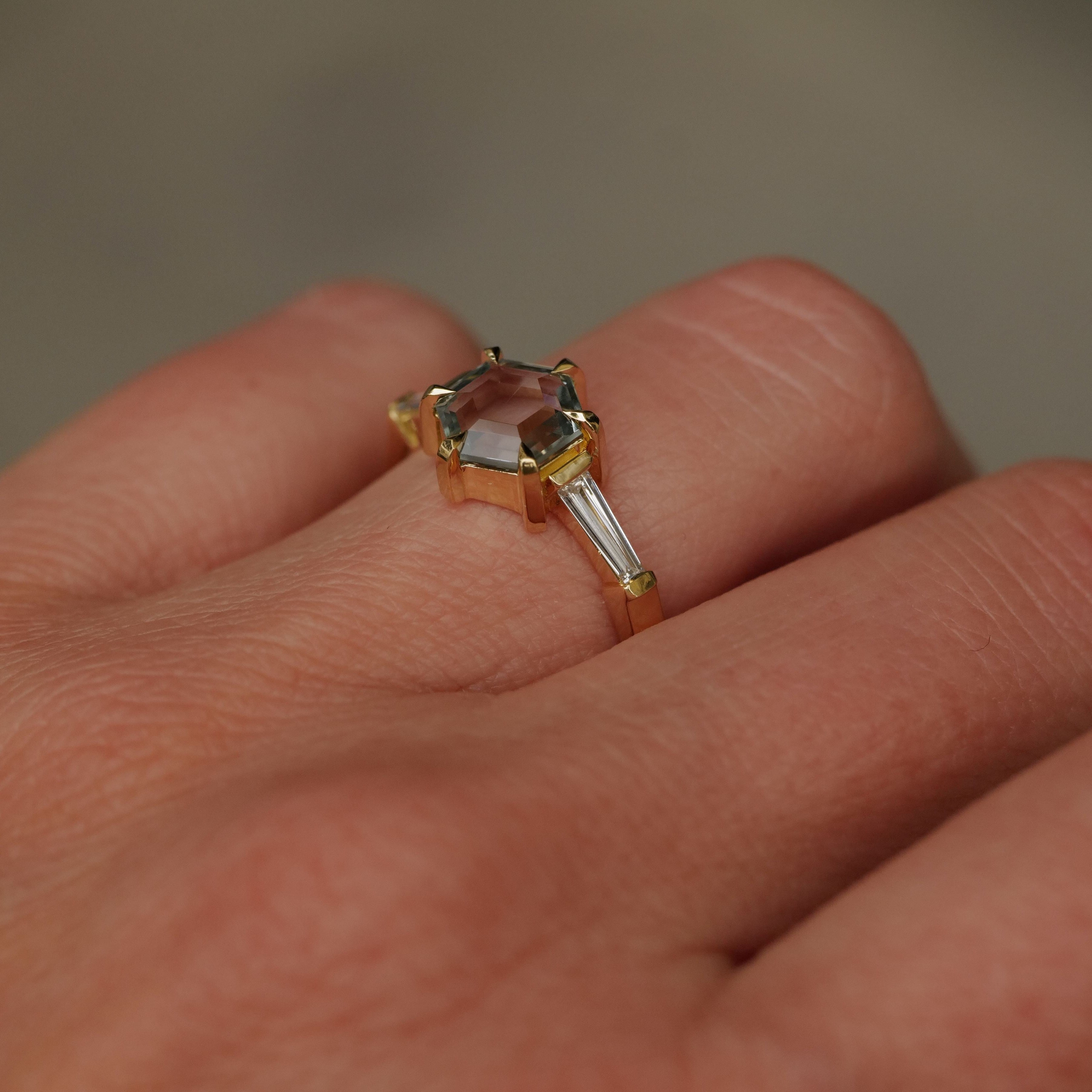 A Fiat Lux Grande Calliope gold ring with a 1.50ct seafoam green hexagon sapphire and baguette diamonds on a finger.