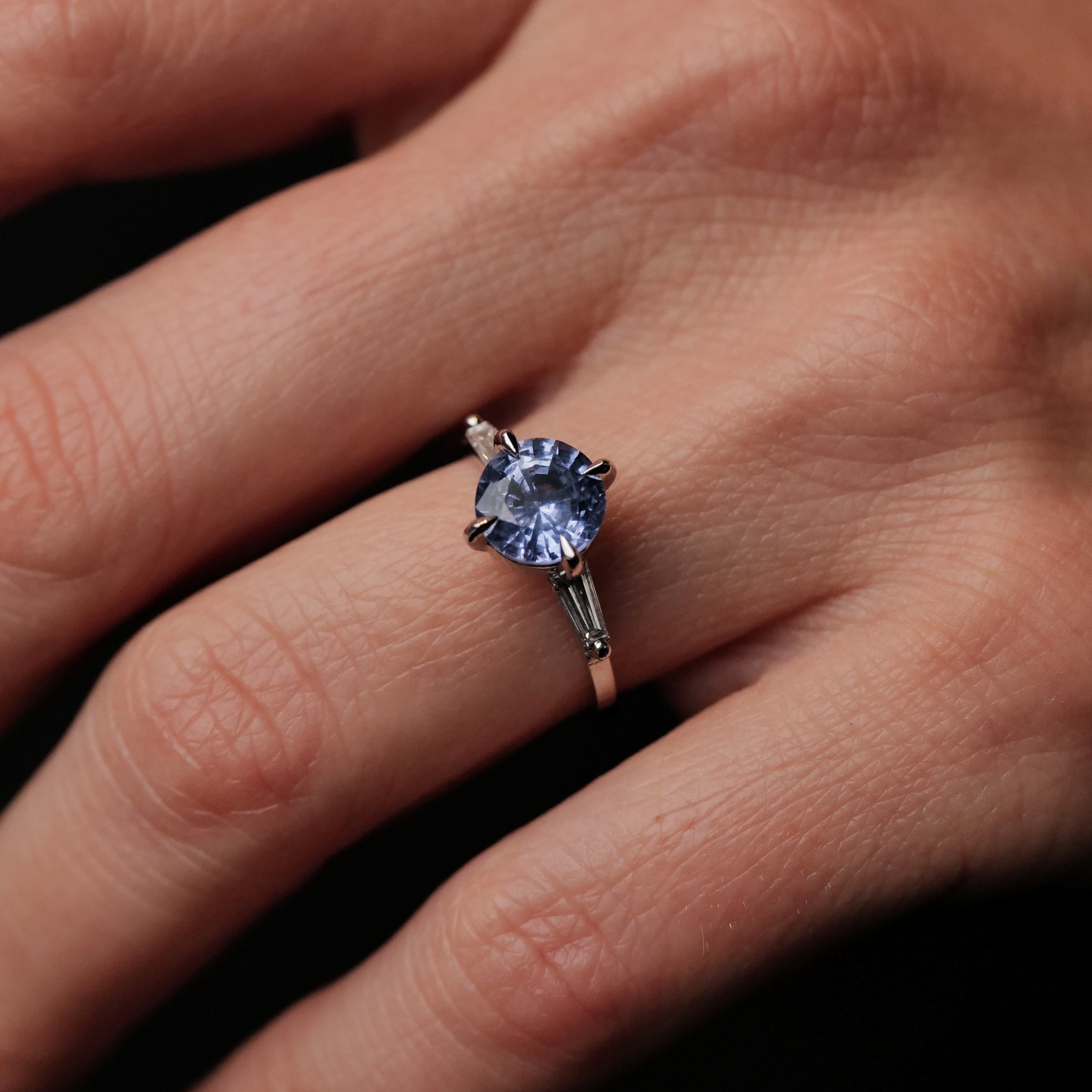 A hand wearing Fiat Luxs Grande Calliope ring with a 2.05ct periwinkle blue sapphire and two clear side stones.