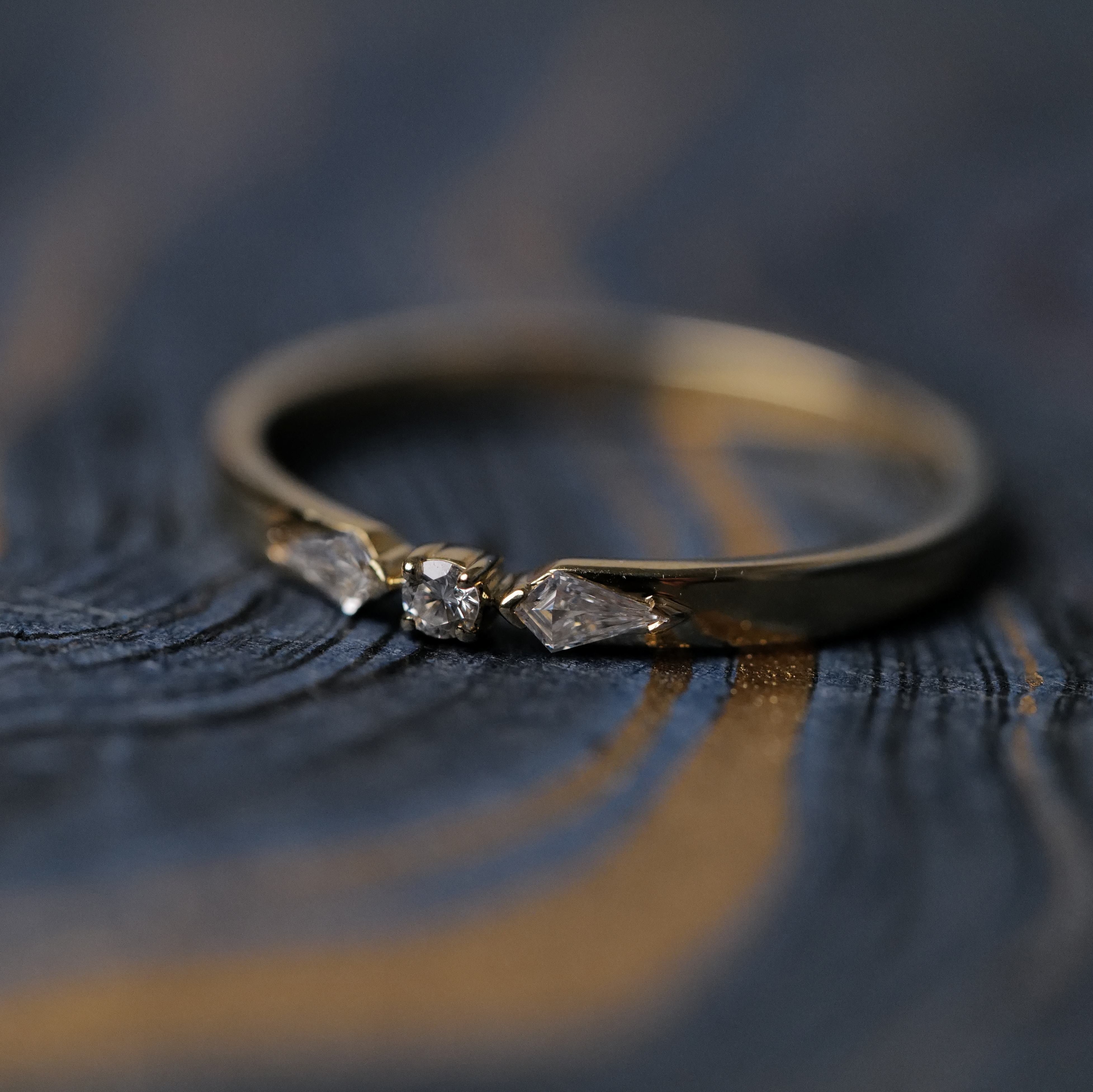Fiat Lux Petite Nova Ring: a 14K gold ring with three diamonds, shown on a dark wood surface with blue and gold streaks.