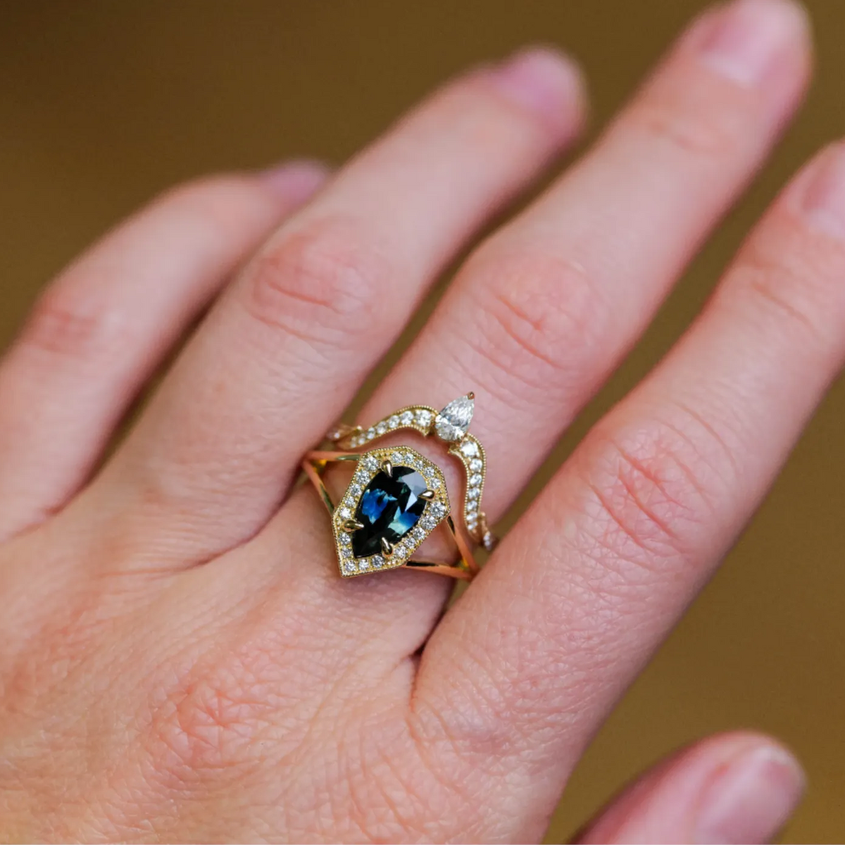 A pear-shaped blue sapphire Dianthe Ring stacked with a diamond-studded chevron band on a finger.