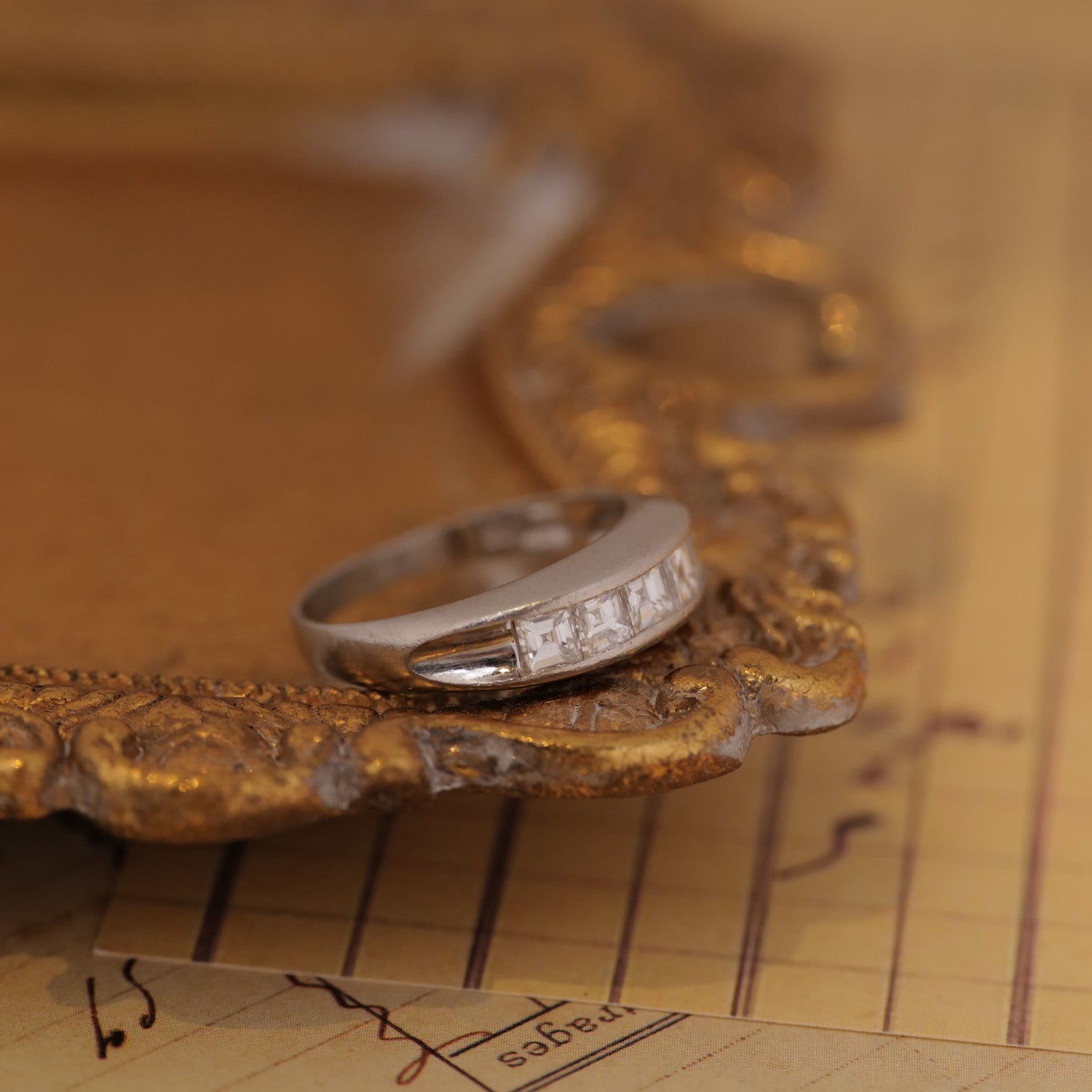 The Vintage Channel Set Carré Diamond Ring is displayed on an ornate gold frame atop handwritten vintage paper.
