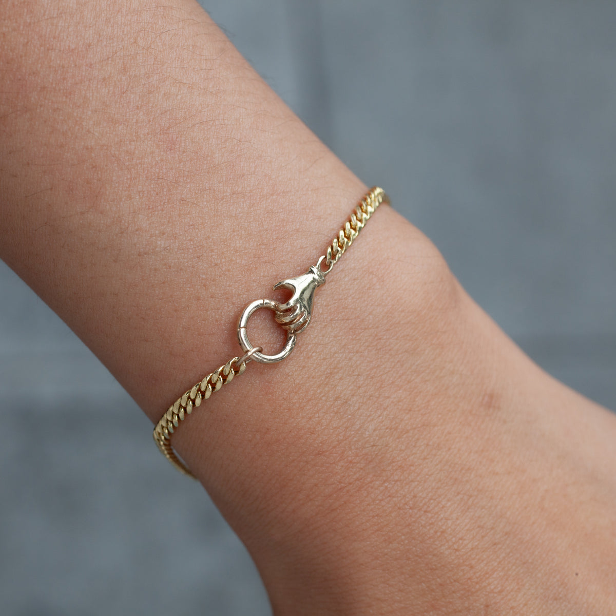 The Acanthus Fob Grip Bracelet, a gold curb chain with a circular clasp, is shown on a wrist with a gray backdrop.
