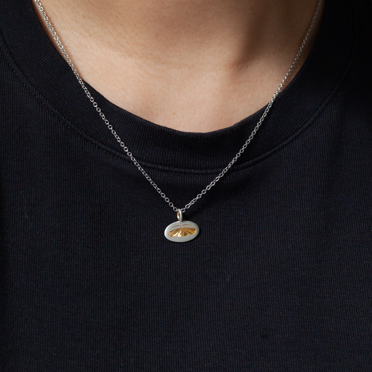 Blue Blue Driver’s Engraved Moth Necklace adds style—oval pendant, gold fan detail, over a black shirt.