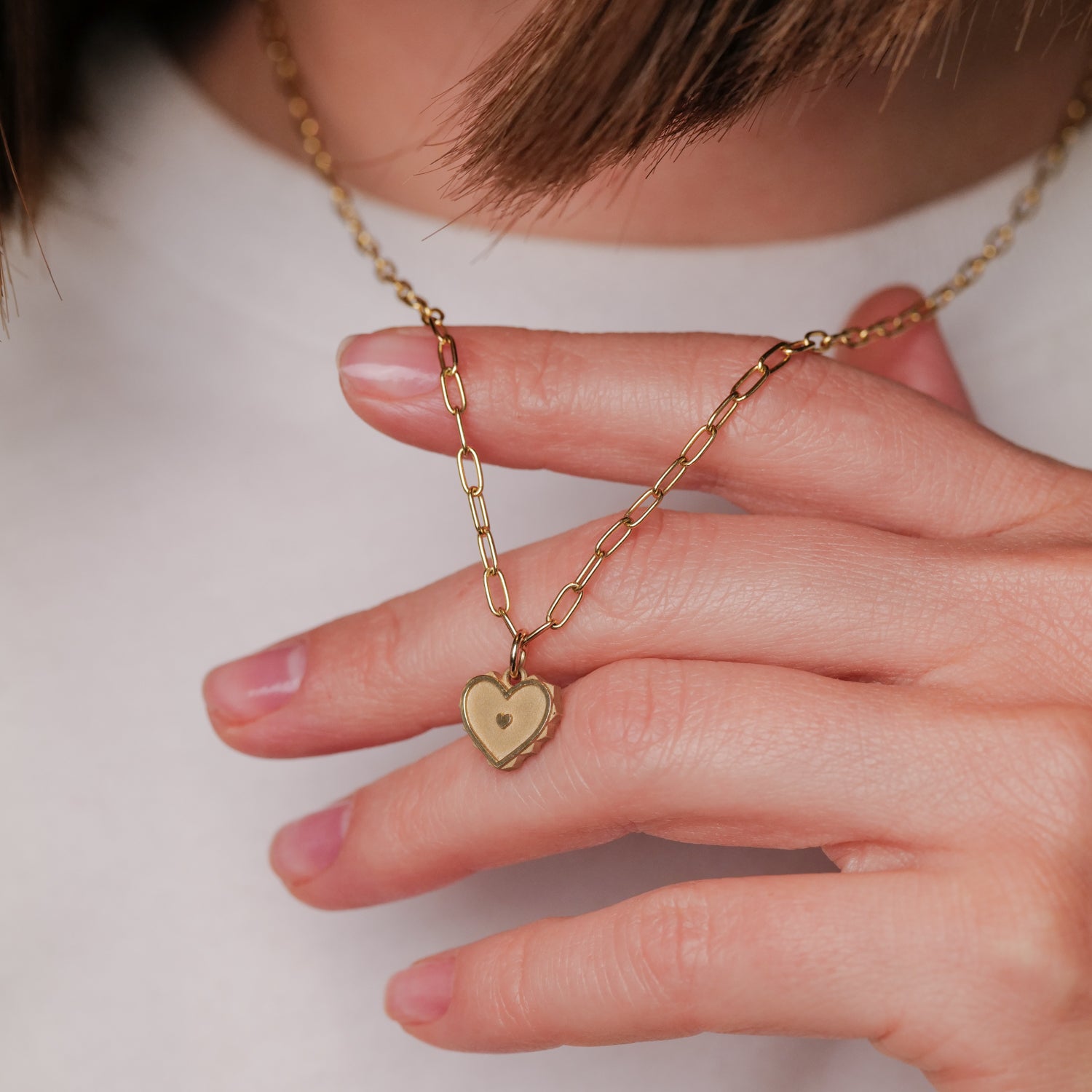 A hand holds EMBLM’s Twin Flame Heart Charm, a 14K gold pendant necklace, worn over a crisp white shirt.