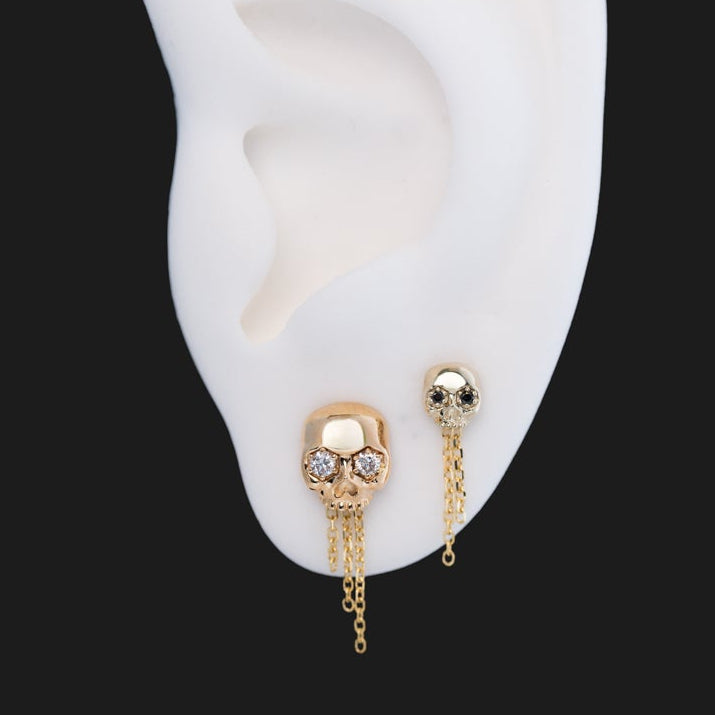 Fiat Lux Three-Chain Dangle earrings: two 14K gold skull studs with dangling chains on a white ear model.